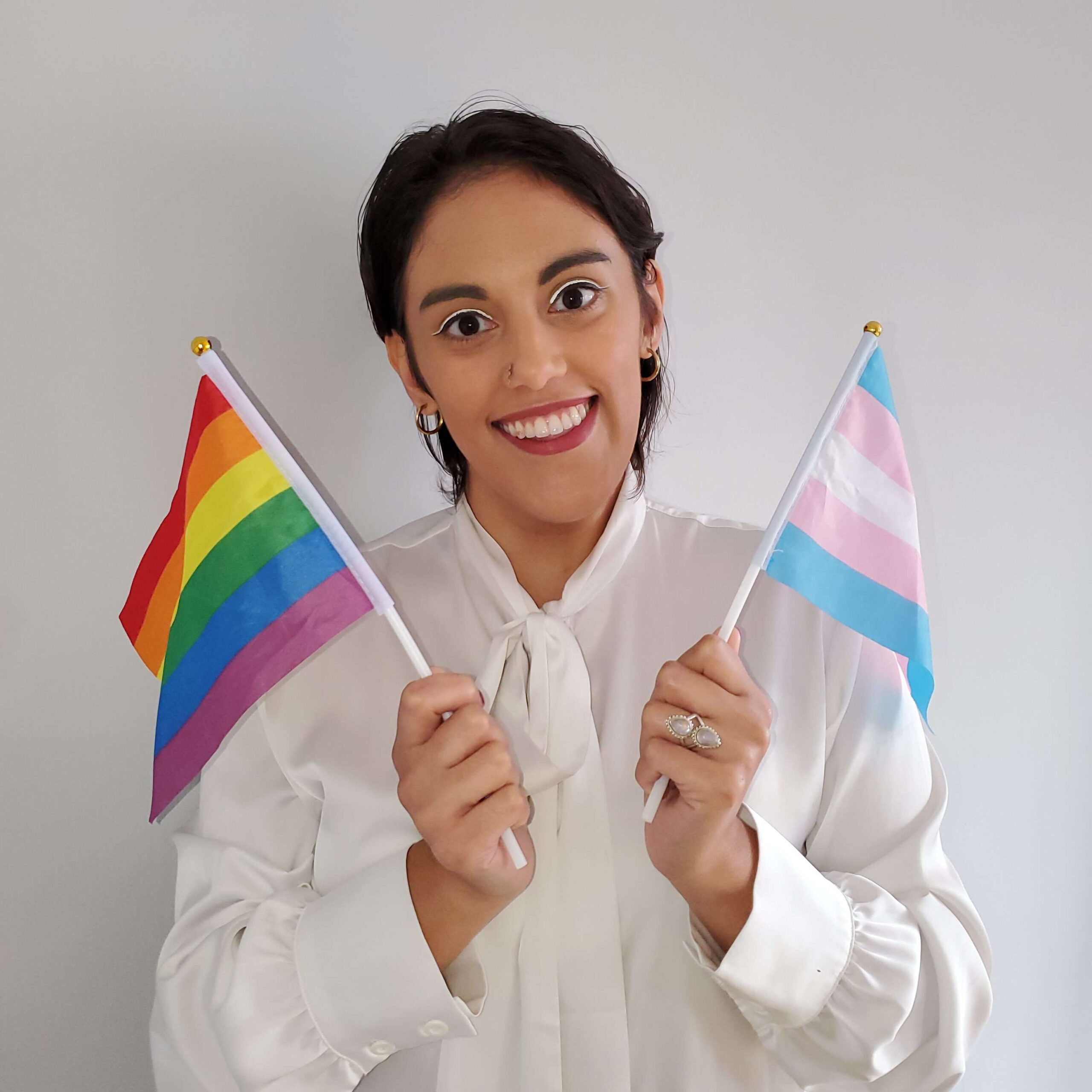 A latine fem person with black hair and red lipstick wears a satin white button up. They're standing against a grey background, holding mini gay and trans flags.