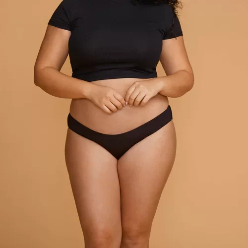 A tan woman stands in the center of an orange background wearing black panties and a black t-shirt