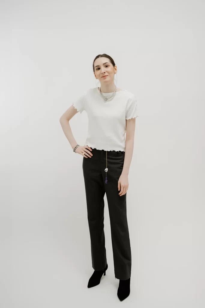 A white woman with pulled back hair stands on a white background with her hand on her hip. She wears a white short-sleeved, lettuce trim shirt and black bootcut pants with a zippered front.