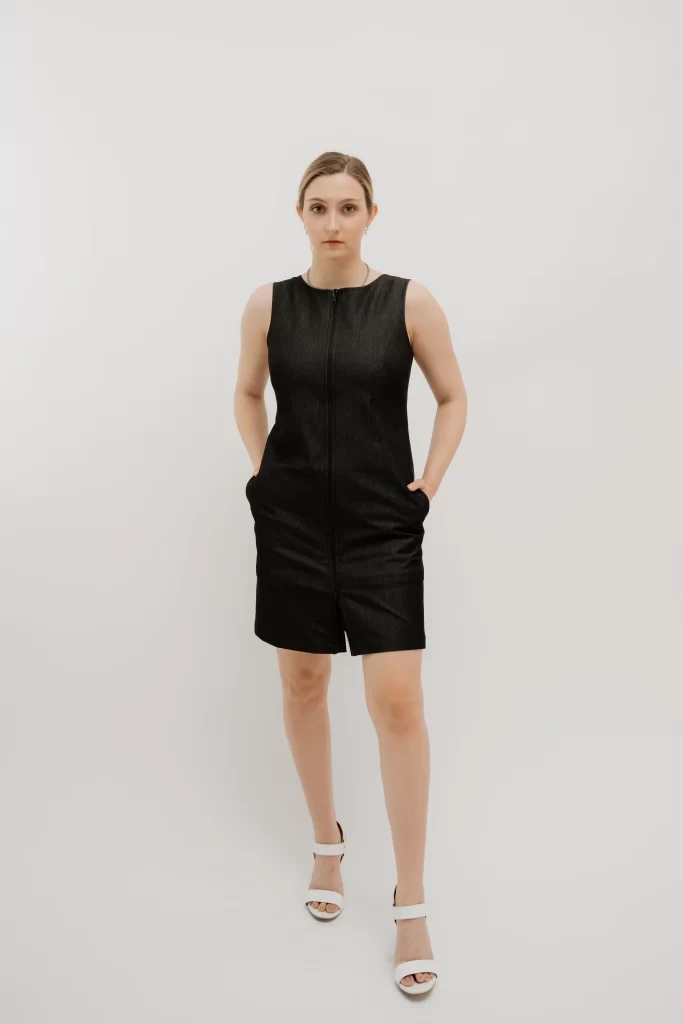 A white woman with her brown hair pulled back stand in front of a white background with her hands in her pockets. She wears a black, boxy, sleeveless dress with zippered front that hits right above the knee.