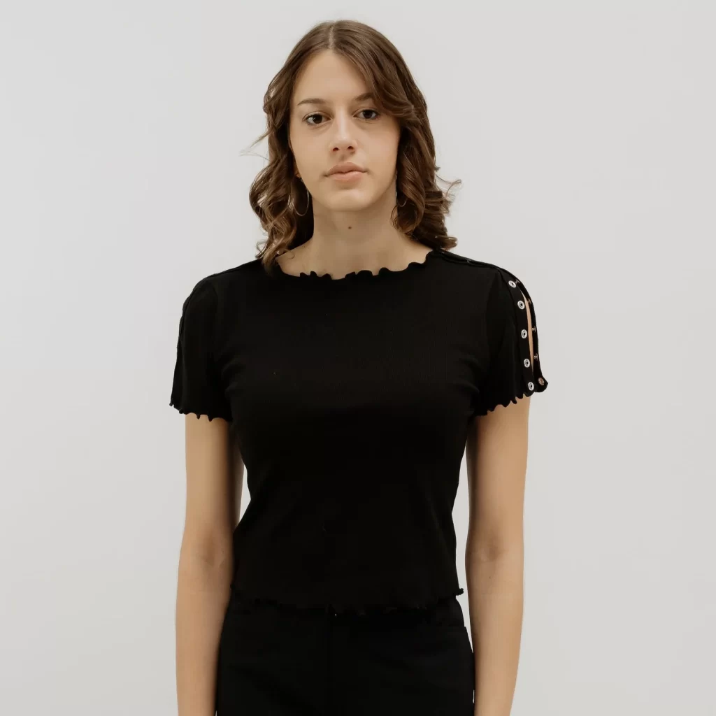 A white woman with brown hair stands in front of a white background. She wears a black, short-sleeve t-shirt with lettuce trim and silver snaps along the shoulders.