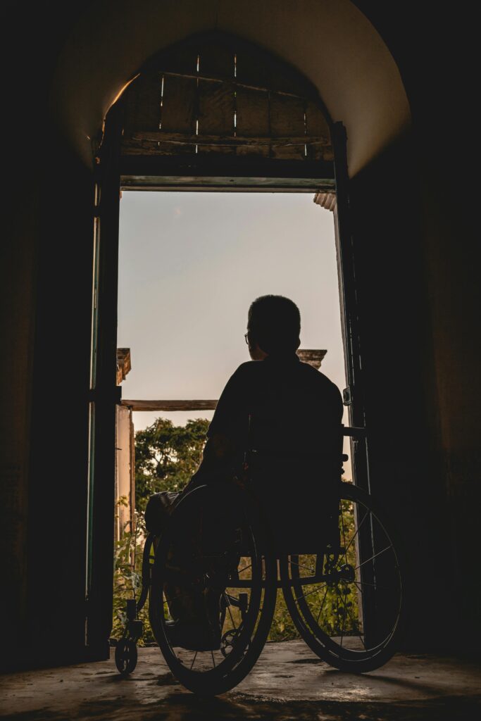 A man in a wheelchair sits in front of an open doorway, looking out over the balcony at the setting sun