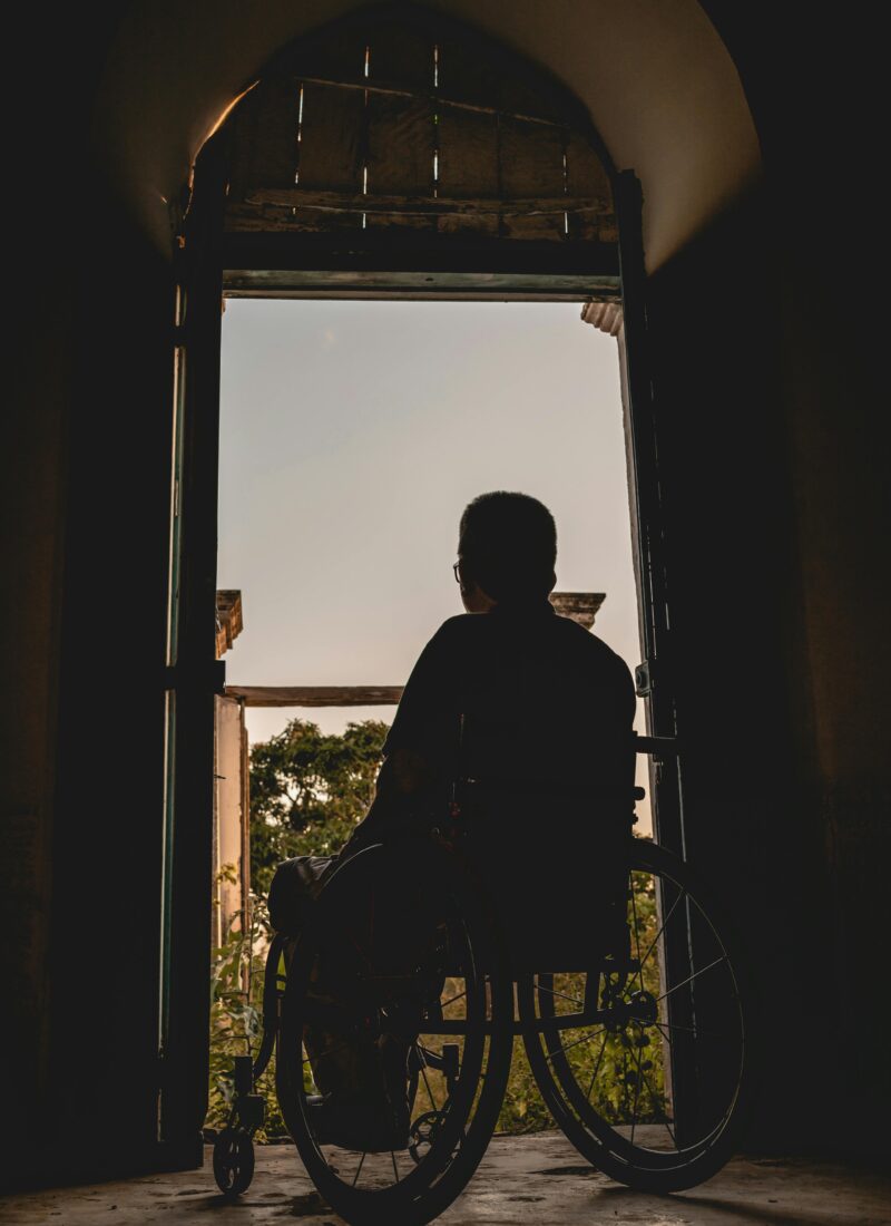 A man in a wheelchair sits in front of an open doorway, looking out over the balcony at the setting sun