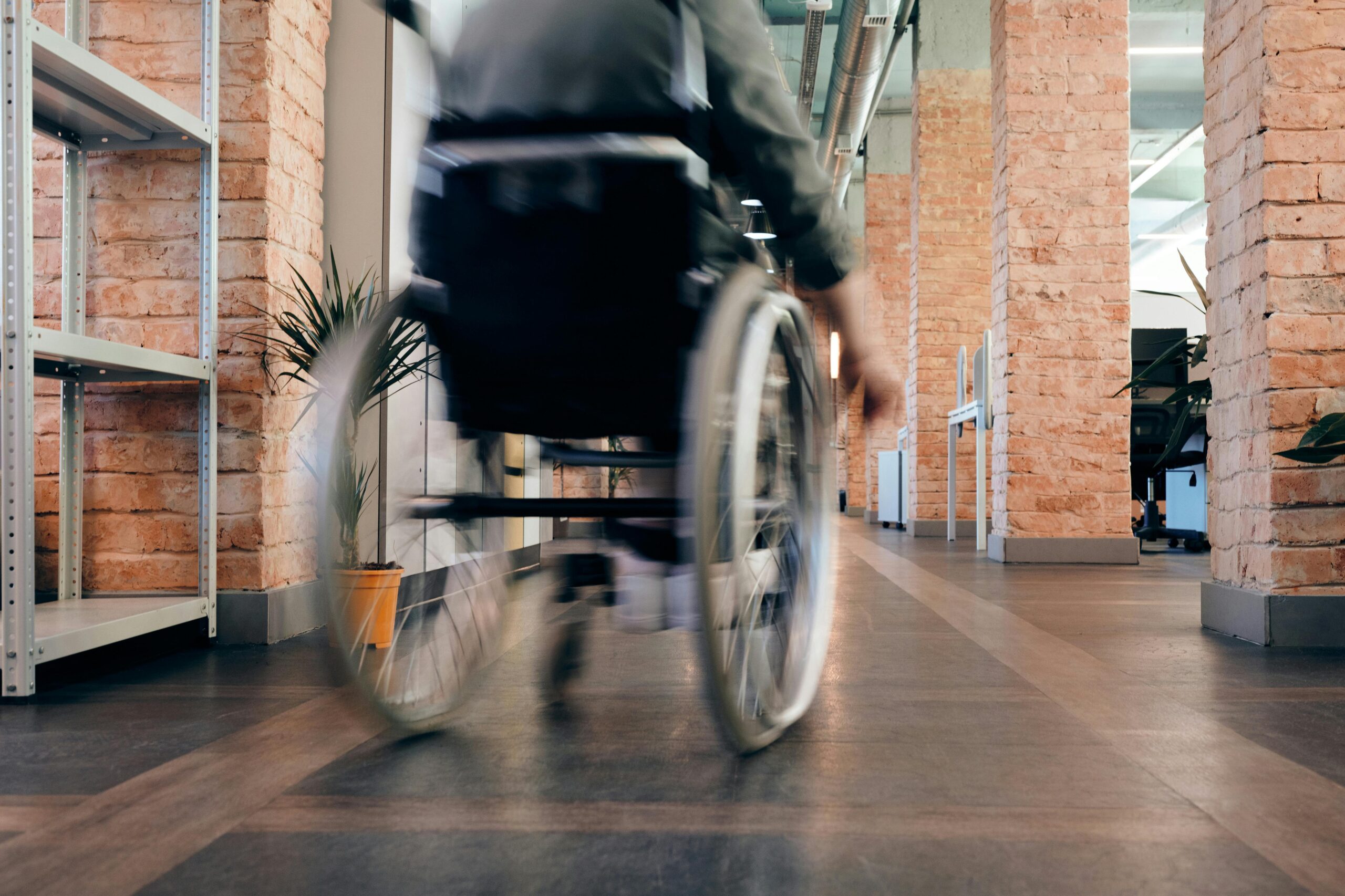 A white person rolls a wheelchair inside a building with black tiles and brick pillars