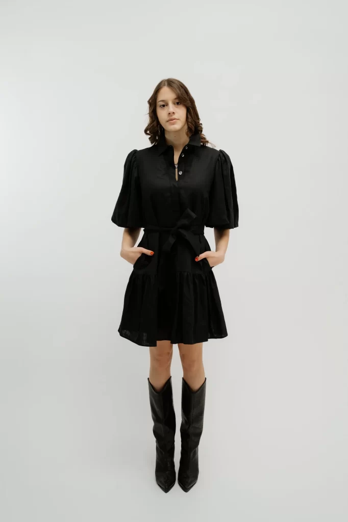 A white woman with brown hair stands in front of a white background. She wears a black dress with sleeves down to her elbows and a button up front. The hem hits right above her knee. She also wears black cowboy boots.